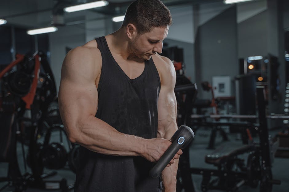 A muscular man uses a massage gun in an indoor gym, focusing on recovery.